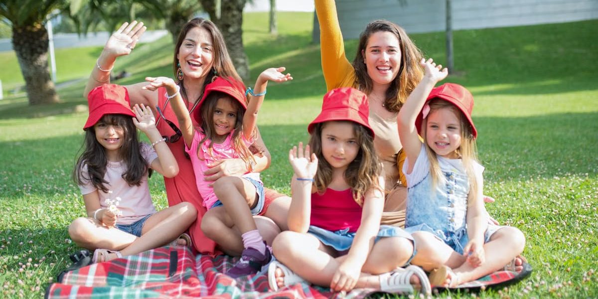 Two women and four girls enjoy a sunny picnic in Portugal, waving happily in the park.