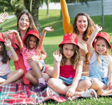 Two women and four girls enjoy a sunny picnic in Portugal, waving happily in the park.